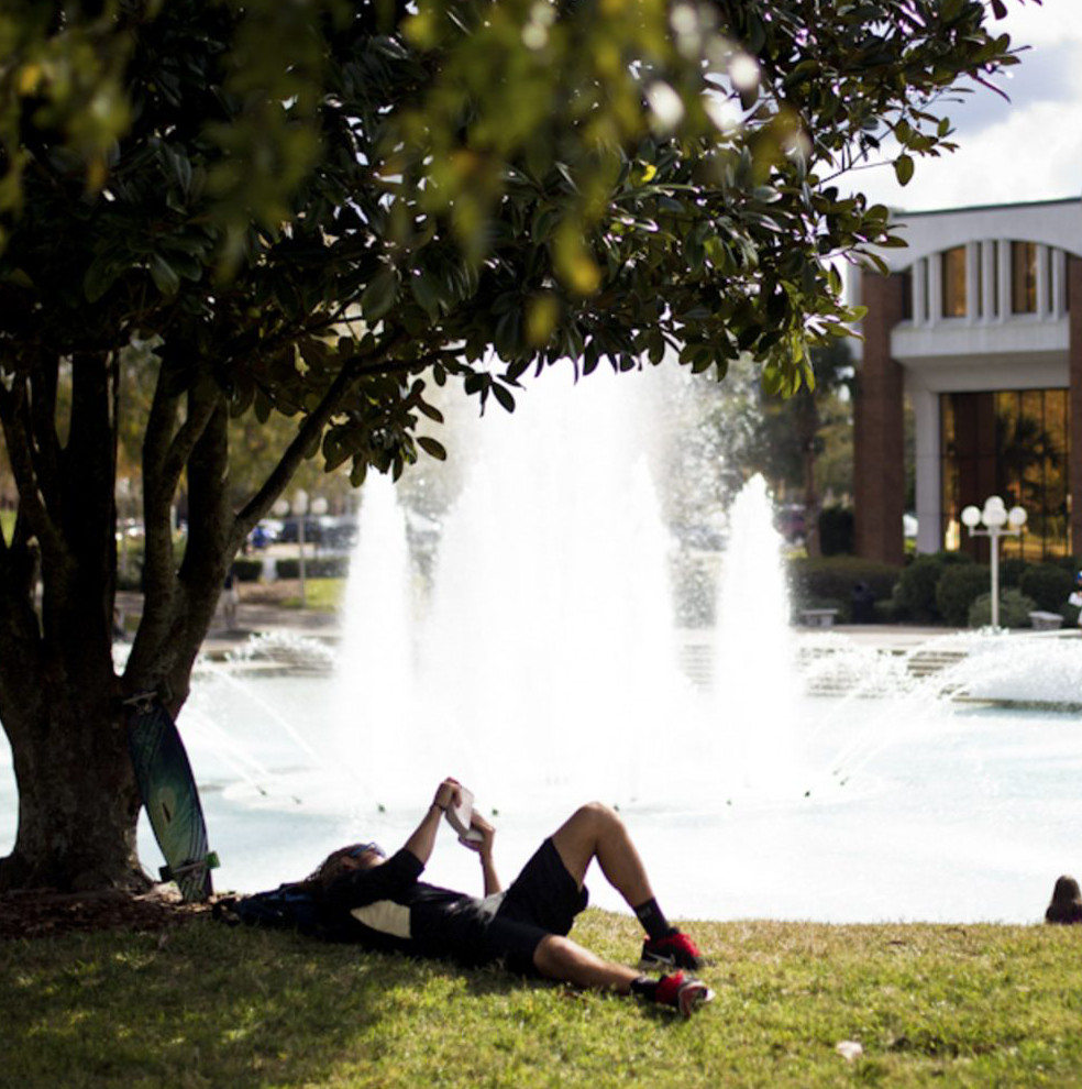UCF student lying down and reading in front of a pond with a fountain on a sunny day.